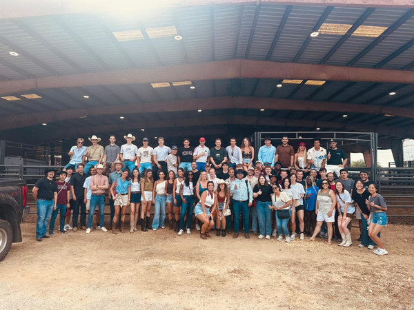 A group of about 30 exchange student at Texas A&M gather together at a rodeo event. The back row of students are sitting on a metal fence while the front row is standing.