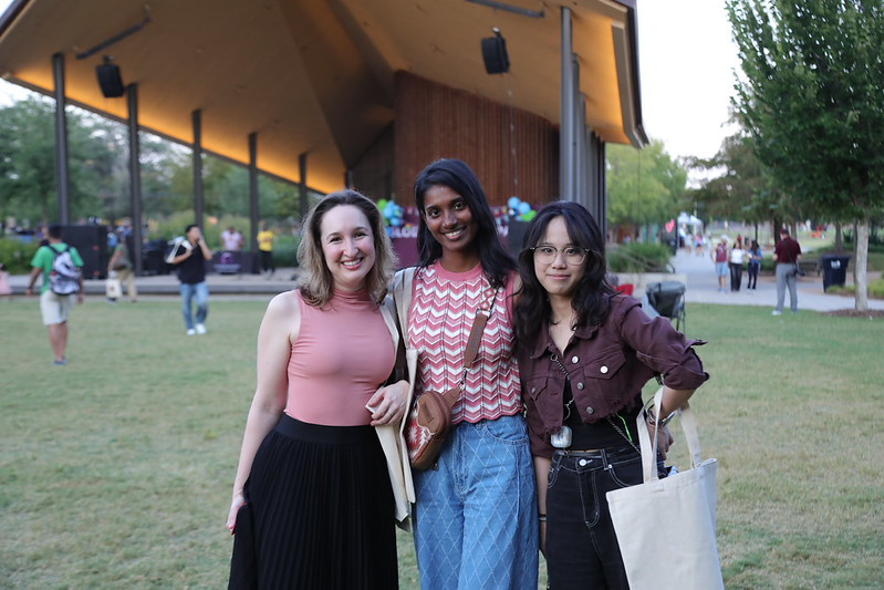 A group of three female Aggies smile for the camera on the field at Aggie Park in front of the stage.