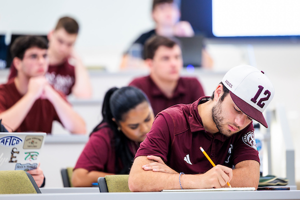 A male student in a college classroom wearing a 12th man hat while looking down and writing on a piece of paper.