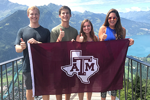 Image of four aggies holding a Texas A&M flag, giving a gig em' in front of a mountain range