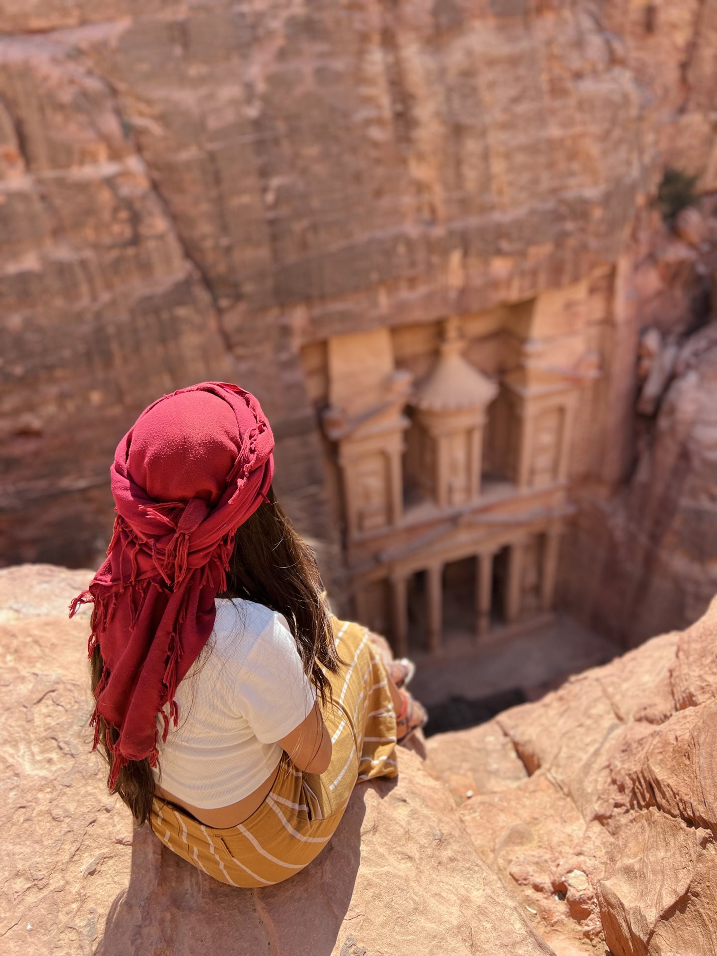 Person sitting on a cliff edge looking down at the entrance to Petra in Jordan